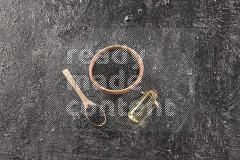 A wooden bowl and spoon full of black seeds and a bottle of black seeds oil on a textured black flooring