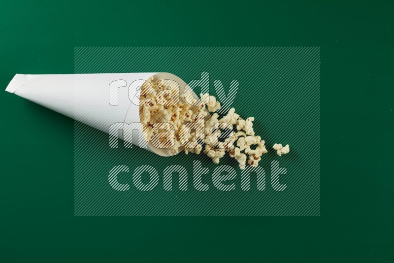 A paper cone full of popcorn and some popcorn came out of it on a green background in a top veiw shot