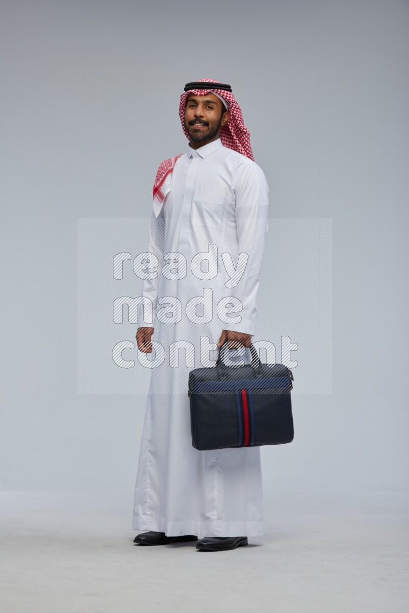 Saudi man Wearing Thob and shomag standing holding bag on Gray background