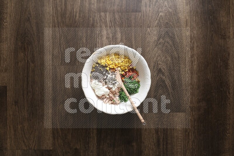 A white bowl full of tuna, colored bell pepper, sour cream, corn, parsley, black pepper powder and sauce, with wooden spoon on wooden background