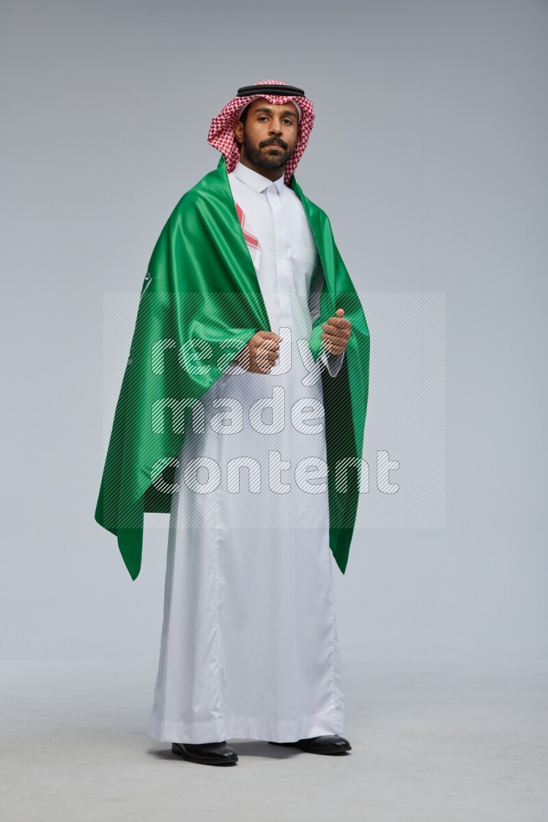 Saudi man Wearing Thob and shomag standing holding Saudi flag on Gray background