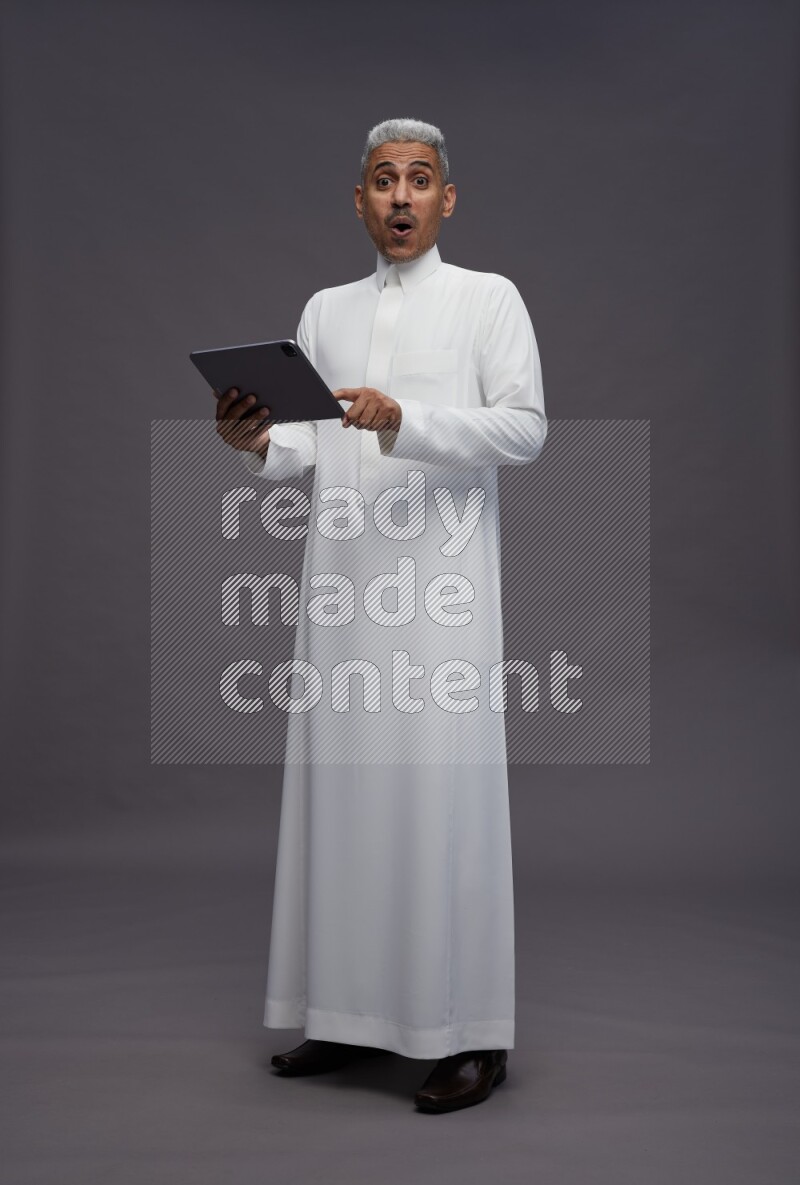 Saudi man wearing thob standing working on tablet on gray background