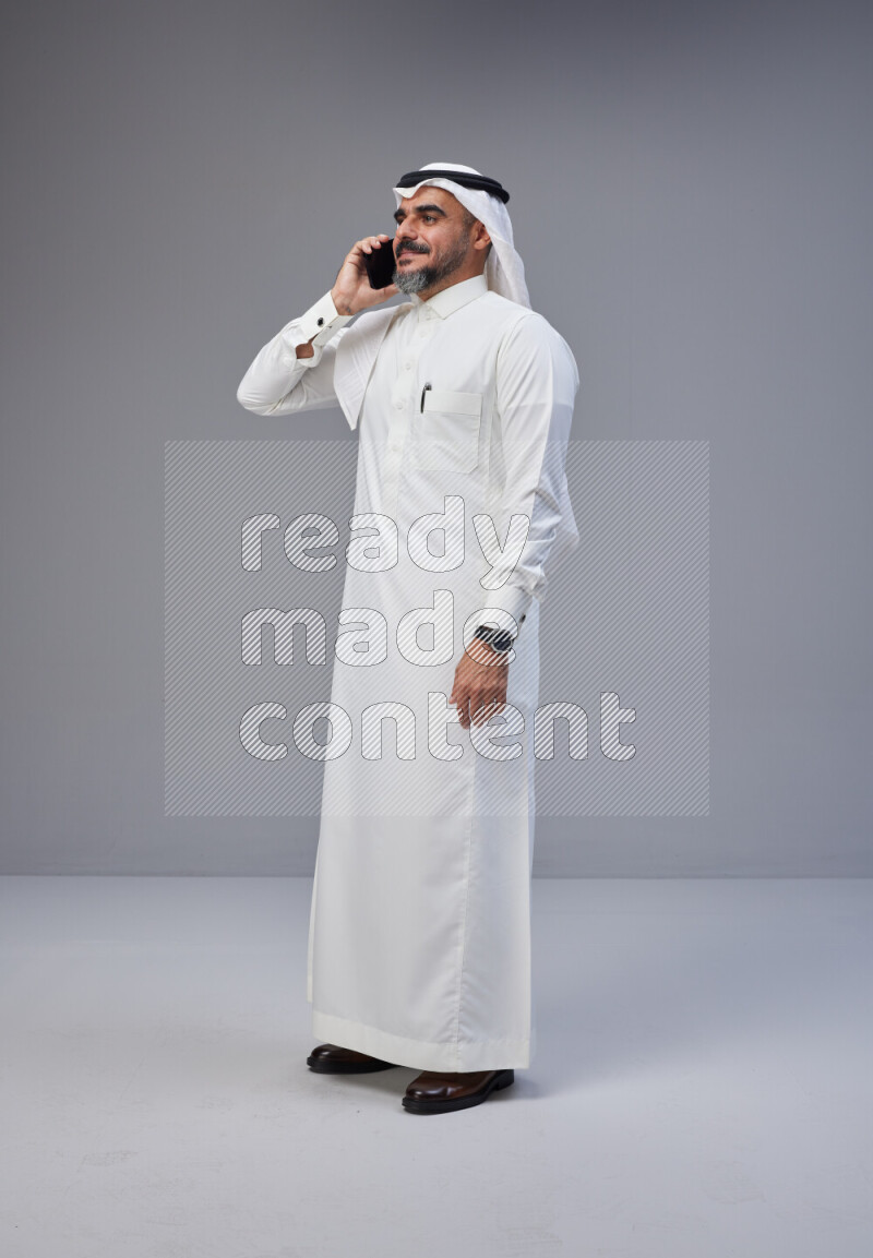Saudi man Wearing Thob and white Shomag standing talking on phone on Gray background