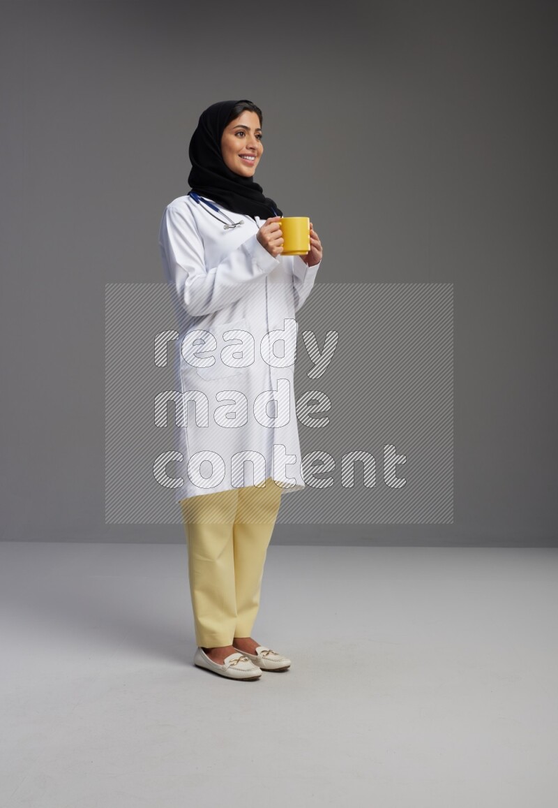 Saudi woman wearing lab coat with stethoscope standing holding mug on Gray background