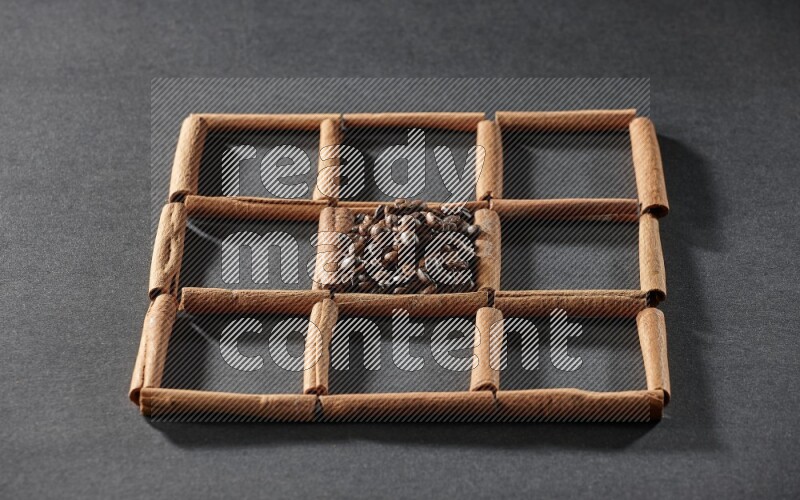 9 squares of cinnamon sticks full of coffee beans in the middle surrounded by nutmeg, dried mint, cloves, dried basil, dried ginger, cinnamon, star anise and cardamom on black flooring