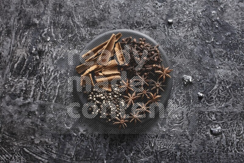 Cinnamon sticks, cloves, star anise and black and white peppers on a black plate on textured black background