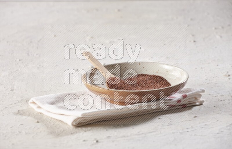 A multicolored pottery plate full of garden cress seeds with a wooden spoon full of the seeds on a napkin on a textured white flooring