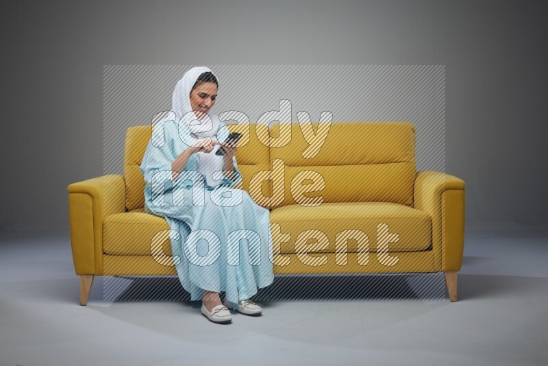 A Saudi woman wearing a light blue Abaya and white head scarf sitting on a yellow sofa and using her phone eye level on a grey background