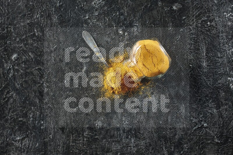 وعاء زجاجي للتوابل مقلوب وملعقة معدنية ممتلئان ببودرة الكركم والبودرة تناثرت منه على خلفية سوداء