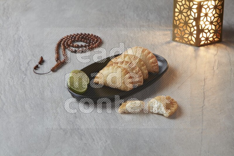 Four fried sambosas in an oval shaped black plate, beside a cut cheese sambosa, a brown misbaha and a golden lantern on a gray background