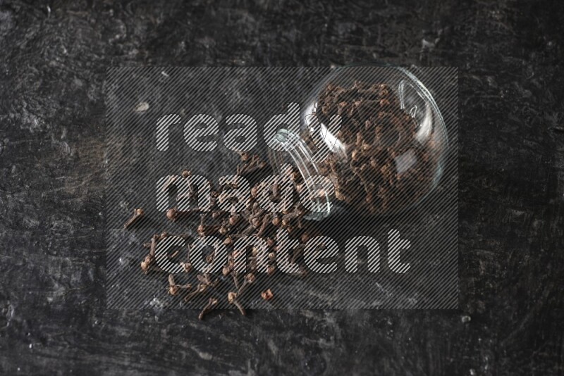 A flipped glass spice jar full of cloves and cloves came out of it on textured black flooring