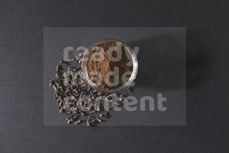 وعاء زجاجي ممتلئ ببودرة القرنفل مع حبوب القرنفل المتناثرة على أرضية سوداء