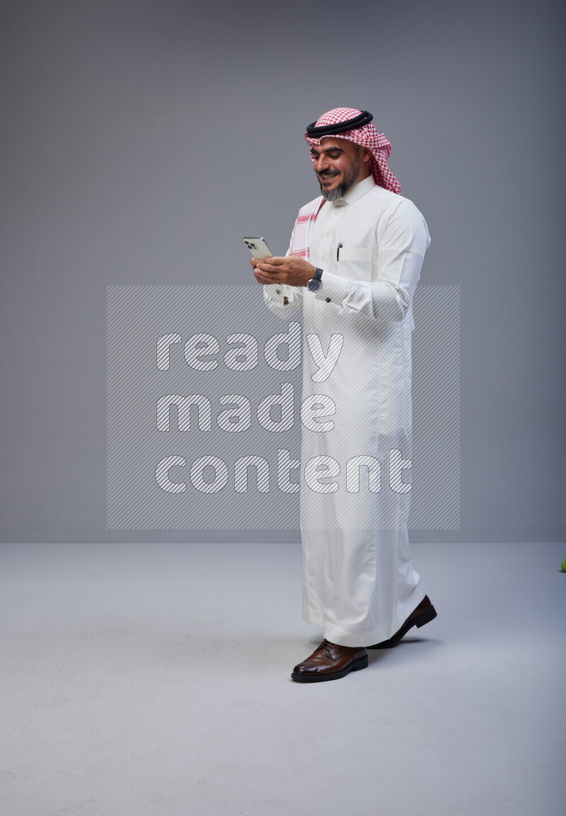 Saudi man Wearing Thob and red Shomag standing texting on phone on Gray background