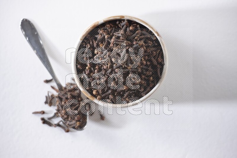 A beige ceramic bowl full of cloves and a metal spoon next to it on a white flooring