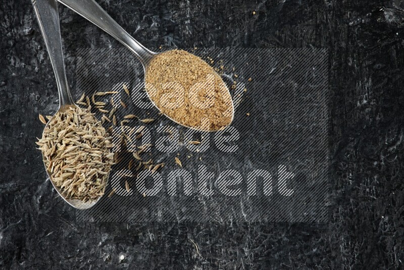 2 metal spoons full of cumin seeds and cumin powder on a textured black flooring