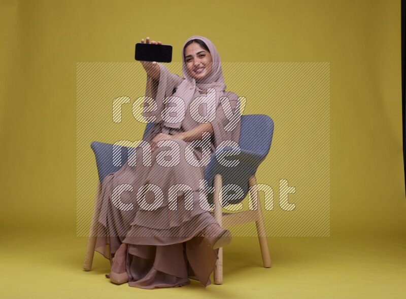 A woman Showing Her Phone Screen Setting  on a Yellow Background wearing Brown Abaya with Hijab