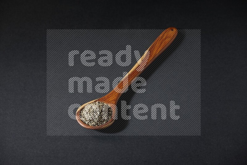 A wooden ladle full of cumin seeds on black flooring