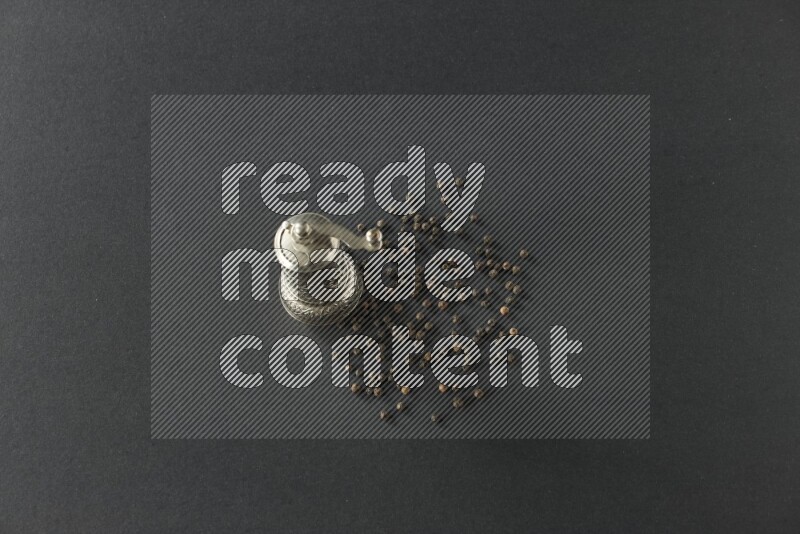 A turkish metal pepper grinder and spreaded black pepper beads on a black flooring