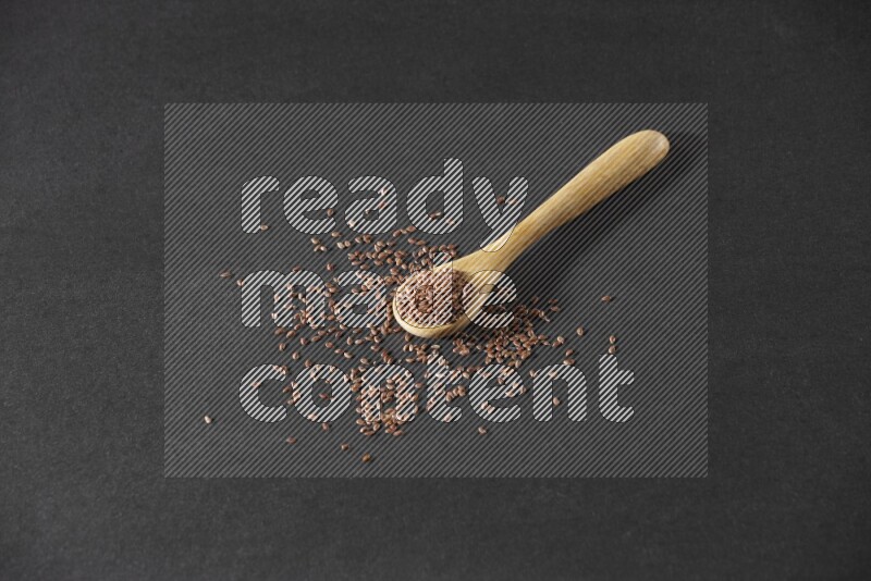 A wooden spoon full of flaxseeds and seeds spread beside it on a black flooring