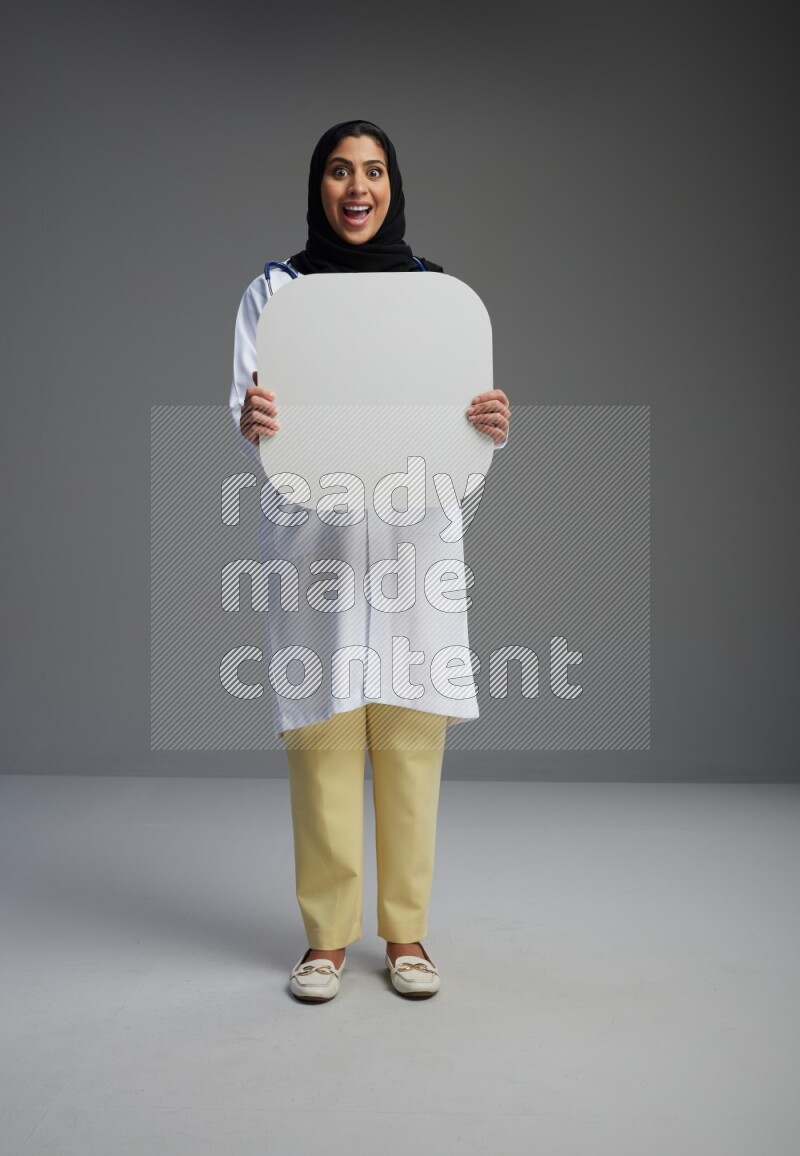 Saudi woman wearing lab coat with stethoscope standing holding social media sign on Gray background