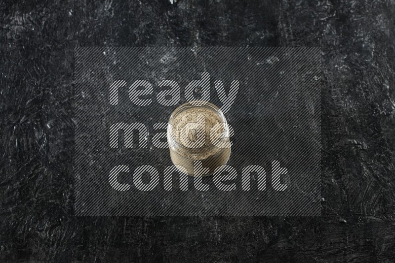 A glass jar full of cardamom powder on textured black flooring
