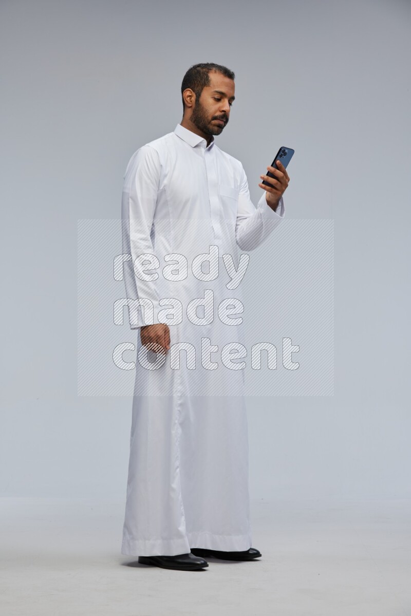 Saudi man wearing Thob standing texting on phone on Gray background