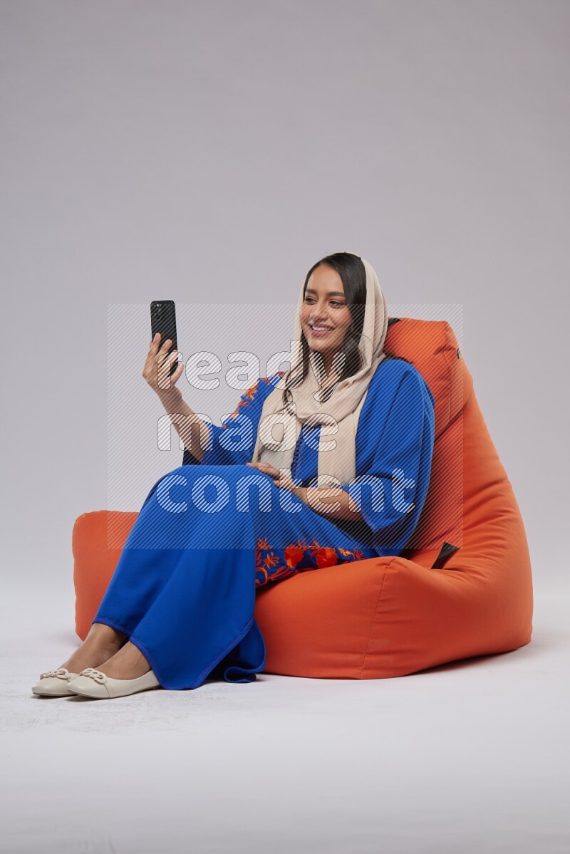 A Saudi woman wearing jalabiya sitting on an orange beanbag and taking selfie