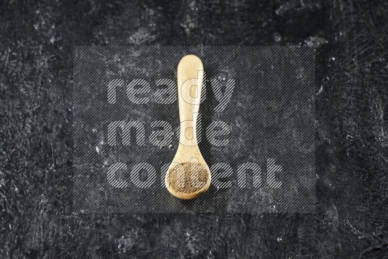 A wooden spoons full of cumin powder on textured black flooring