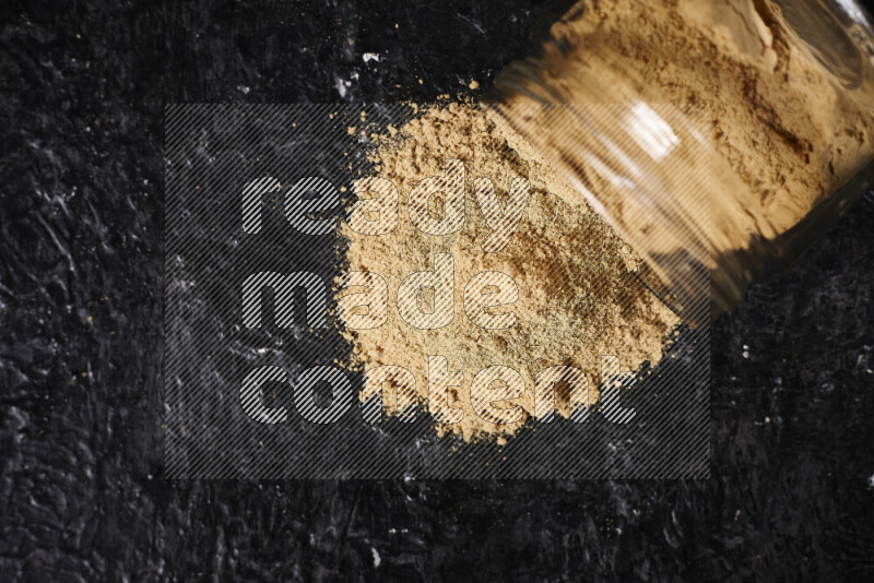 A glass jar full of ground ginger powder flipped with some spilling powder on black background