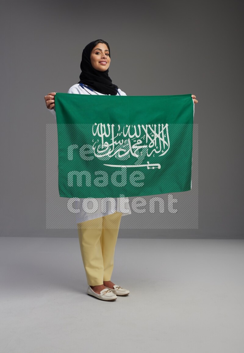 Saudi woman wearing lab coat with stethoscope standing holding Saudi flag on Gray background