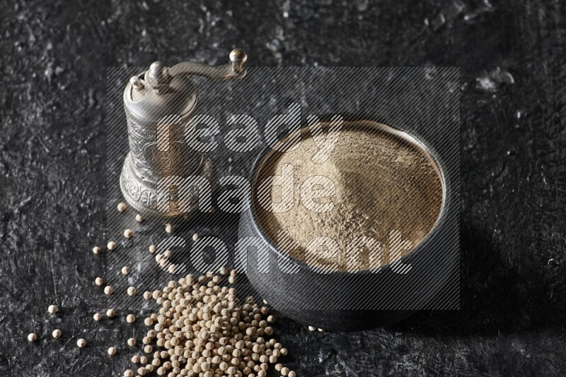 Black pottery bowl full of white pepper powder with pepper beads and metal pepper grinder on textured black flooring