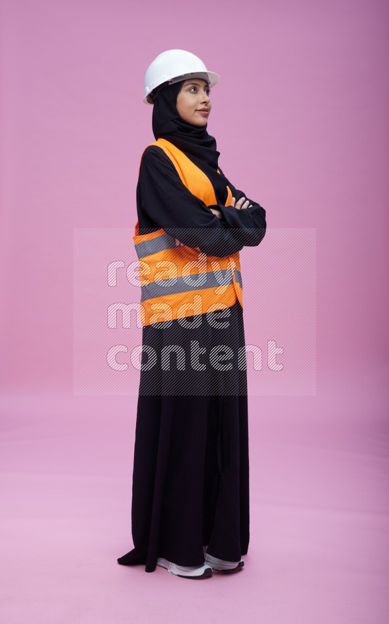 Saudi woman wearing Abaya with engineer vest and helmet standing with crossed arms on pink background