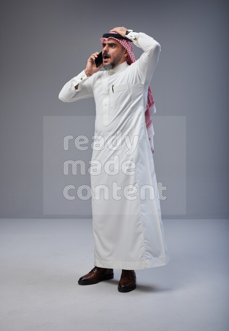 Saudi man Wearing Thob and red Shomag standing talking on phone on Gray background