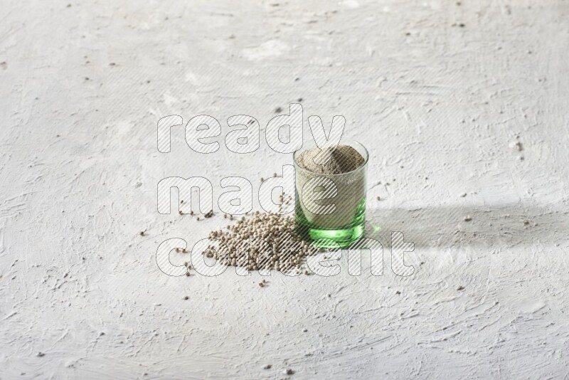 A green glass cup full of white pepper powder with white pepper beads on textured white flooring