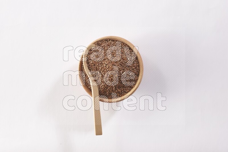 A wooden bowl full of flax seeds and a wooden spoon full of flax seeds in it on a white flooring