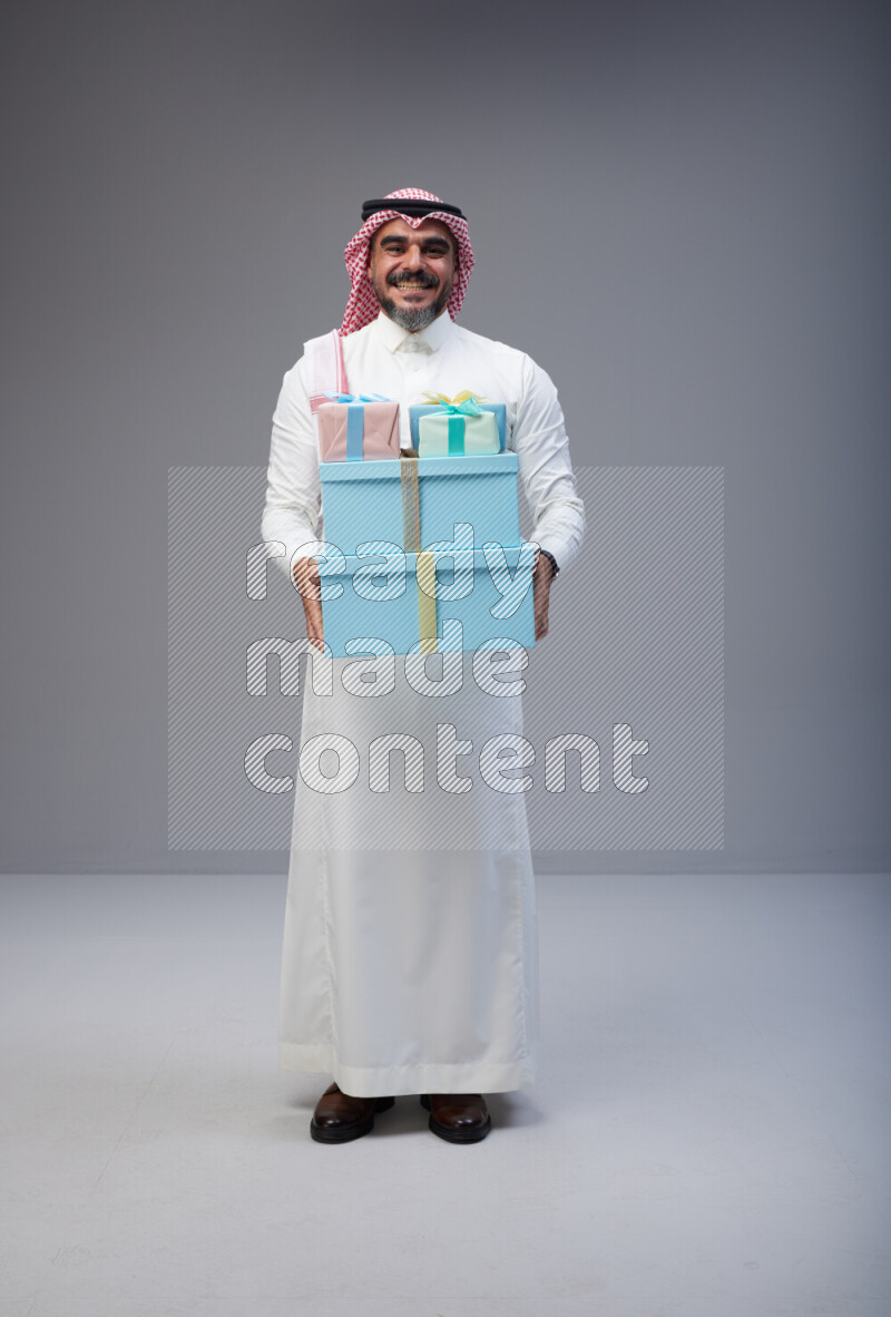 Saudi man Wearing Thob and red Shomag standing holding gift box on Gray background
