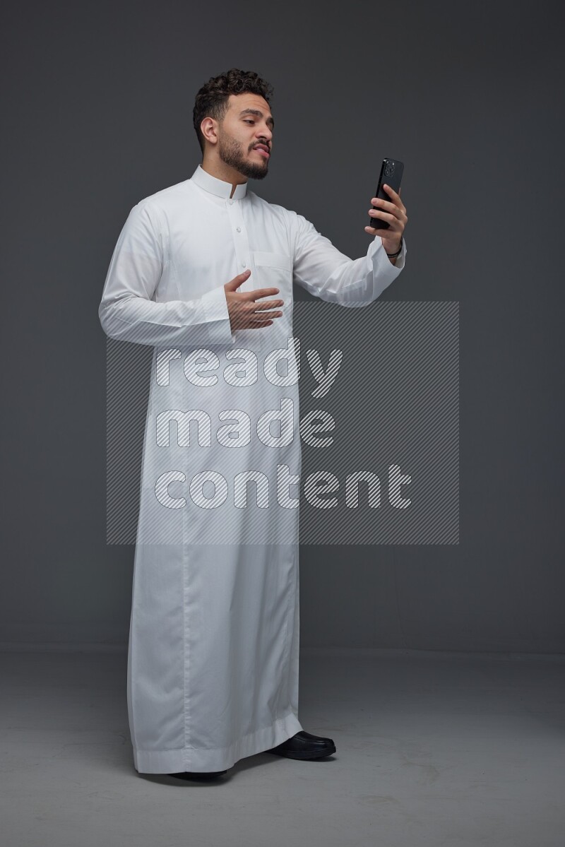 A Saudi man wearing Thobe and taking selfie with his phone making different poses eye level on a gray background