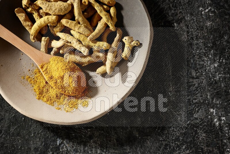 A plate filled with dried turmeric fingers and a wooden spoon full of turmeric powder on a textured black flooring