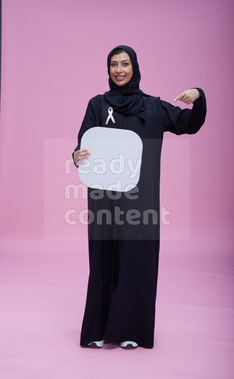 Saudi woman wearing pink ribbon on Abaya standing holding social media signs on pink background