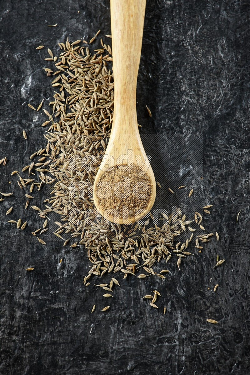 A wooden spoon full of cumin powder and cumin seeds spreaded on textured black flooring
