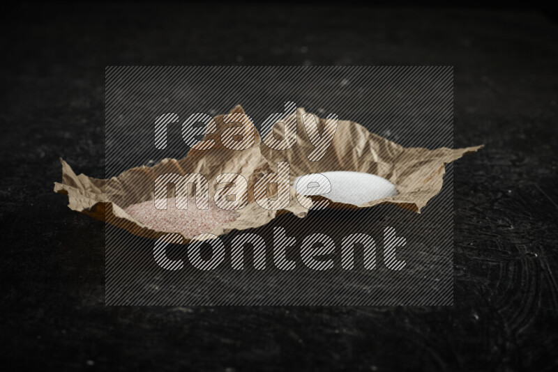2 crumpled pieces of paper full of pink himalayan salt and white salt on black background