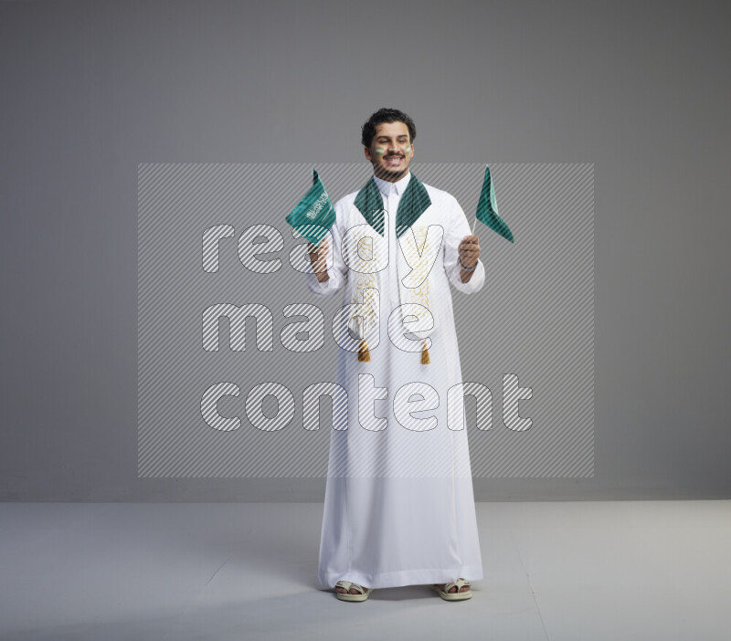 A Saudi man standing wearing thob and saudi flag scarf with face painting holding small Saudi flag on gray background