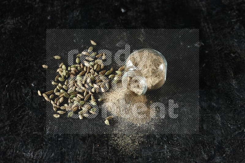 وعاء زجاجي للتوابل مقلوب وممتلئ ببودرة الهيل والبودرة تناثرت منه مع حبوب الهيل المتناثرة على خلفية سوداء