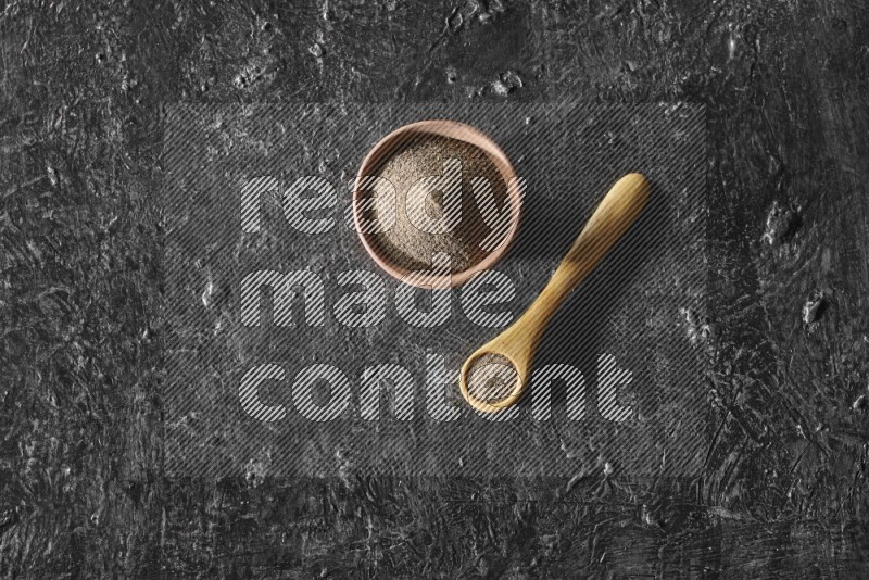 A wooden bowl and wooden spoon full of black pepper powder on a textured black flooring