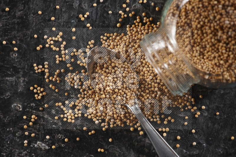 A glass spice jar and a metal spoon full of mustard seeds and jar is flipped with fallen seeds on a textured black flooring