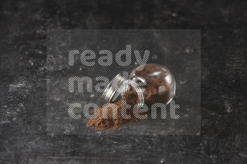 A flipped glass spice jar full of cloves powder and powder came out of it on textured black flooring