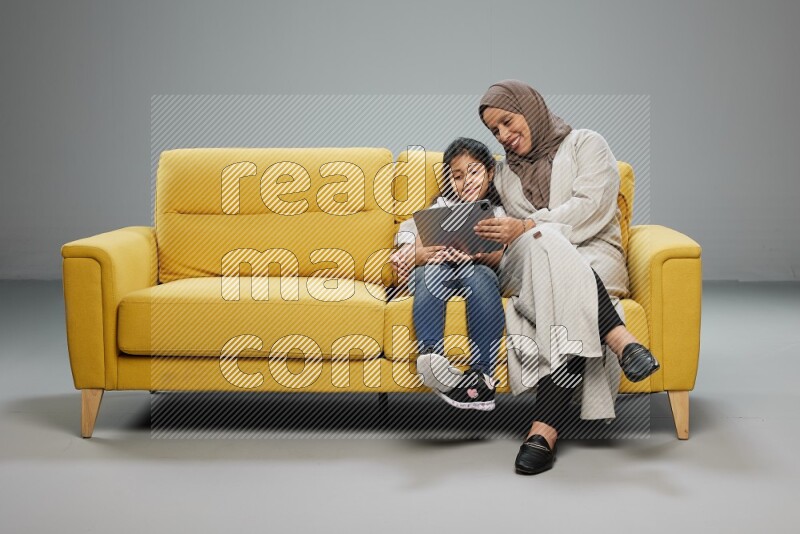 A girl with her mother on yellow sofa and watching on iPad on gray background