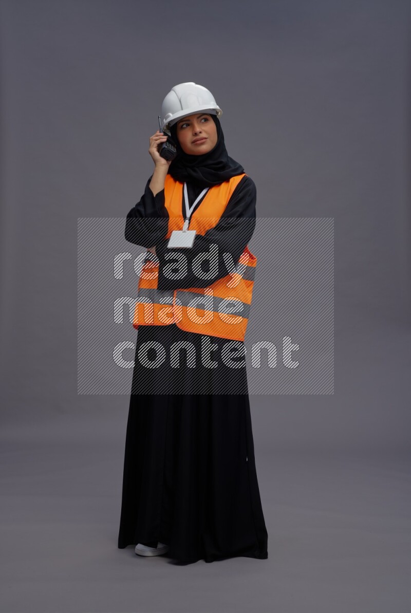 Saudi woman wearing Abaya with engineer vest with neck strap employee badge standing holding walkie-talkie on gray background