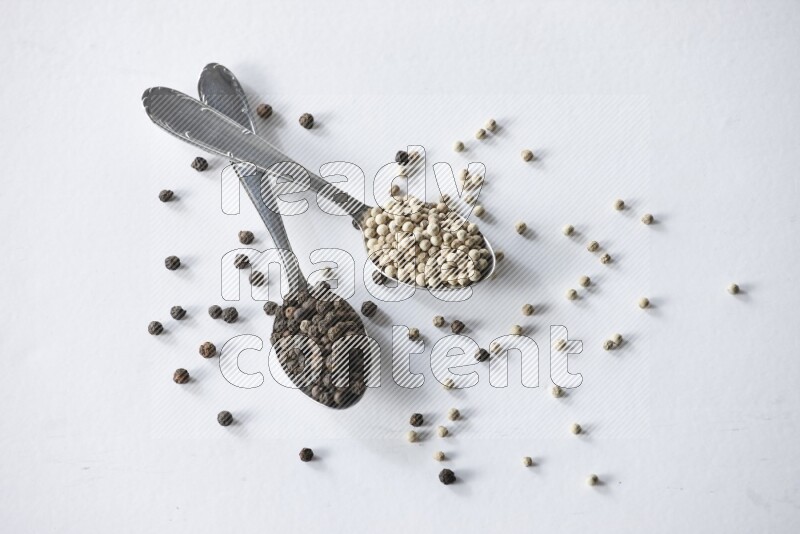 2 metal spoons full of black and white pepper beads with spreaded beads on white flooring