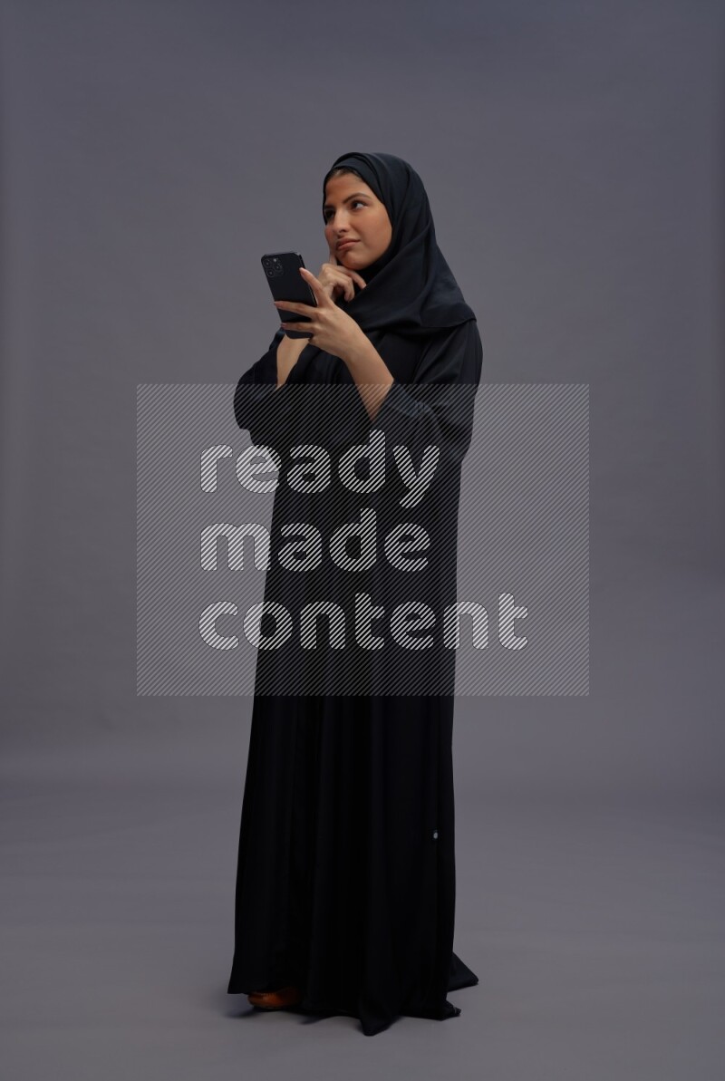 Saudi woman wearing Abaya standing texting on phone on gray background